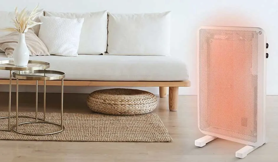 Een lichte woonkamer toont een witte zetel met kussens, gouden tafels, een rieten poef en een witte radiator. Een jute tapijt ligt op de houten vloer.