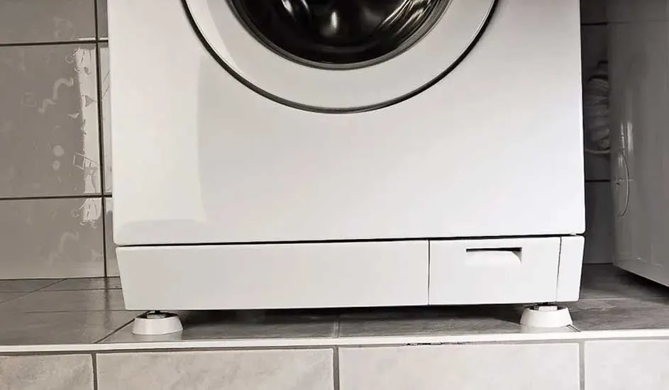 Le bas d’un lave-linge blanc sur du carrelage, des pieds amortisseurs sous les pieds, la marque n’est pas visible sur l’appareil.