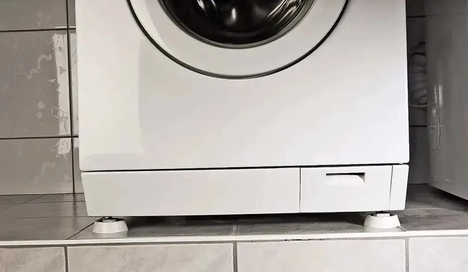 De onderkant van een witte wasmachine op tegels, met dempende voeten onder de poten, merk is niet zichtbaar op de machine.