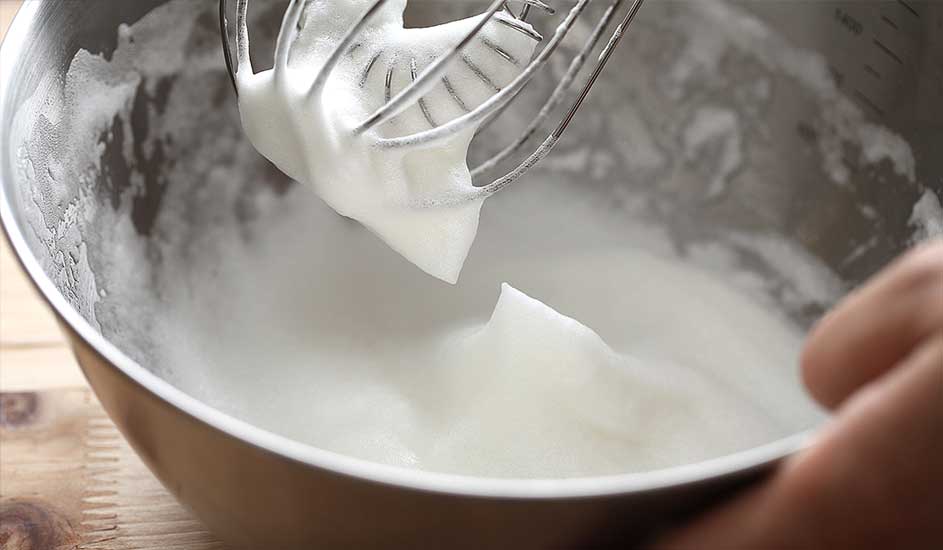 Un bol en métal contient des blancs d'œufs battus, un fouet métallique est au-dessus.