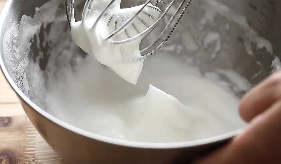 Un bol en métal contient des blancs d'œufs battus, un fouet métallique est au-dessus.