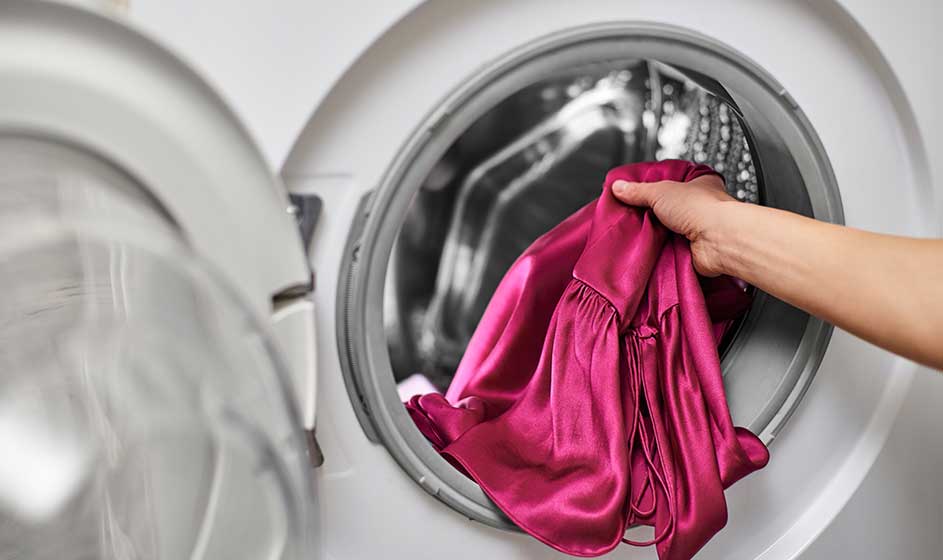 Een hand stopt een glanzend roze kledingstuk in de geopende trommel van een wasmachine, klaar om zorgvuldig gewassen te worden.
