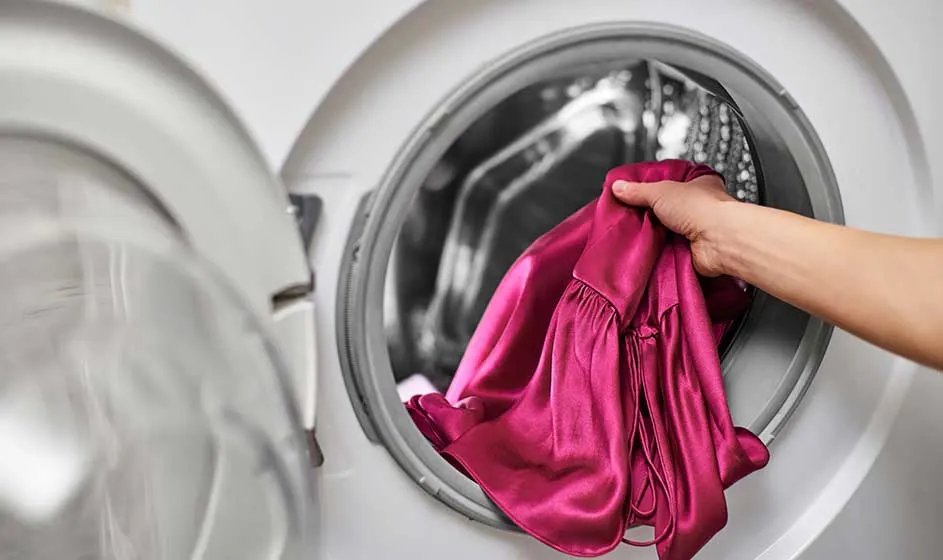 Een hand stopt een glanzend roze kledingstuk in de geopende trommel van een wasmachine, klaar om zorgvuldig gewassen te worden.