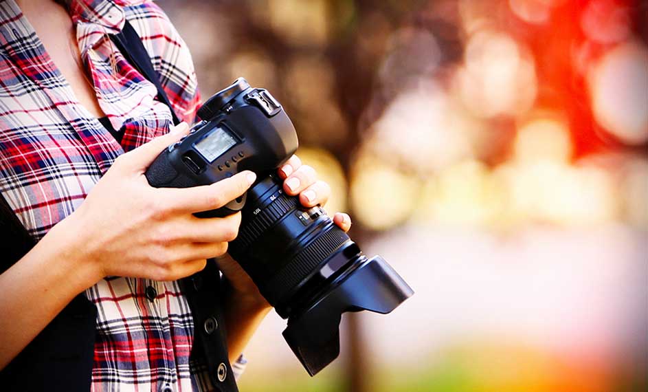 Een zwarte camera met een lange lens wordt vastgehouden tegen een vage achtergrond van groene en oranje tinten.