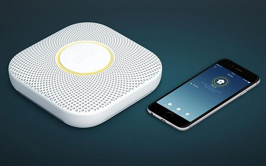 Un détecteur de fumée blanc à grille se trouve à côté d'un smartphone sur fond bleu. L'écran affiche une application.