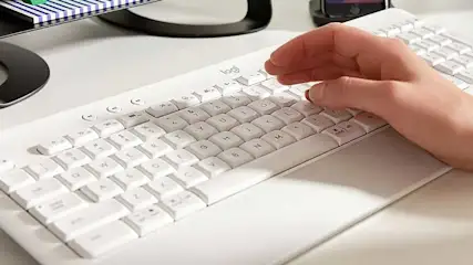 Een witte Logitech toetsenbord ligt op een bureau, met een hand die een toets indrukt. Achtergrond: monitorvoet, bureau-accessoires.