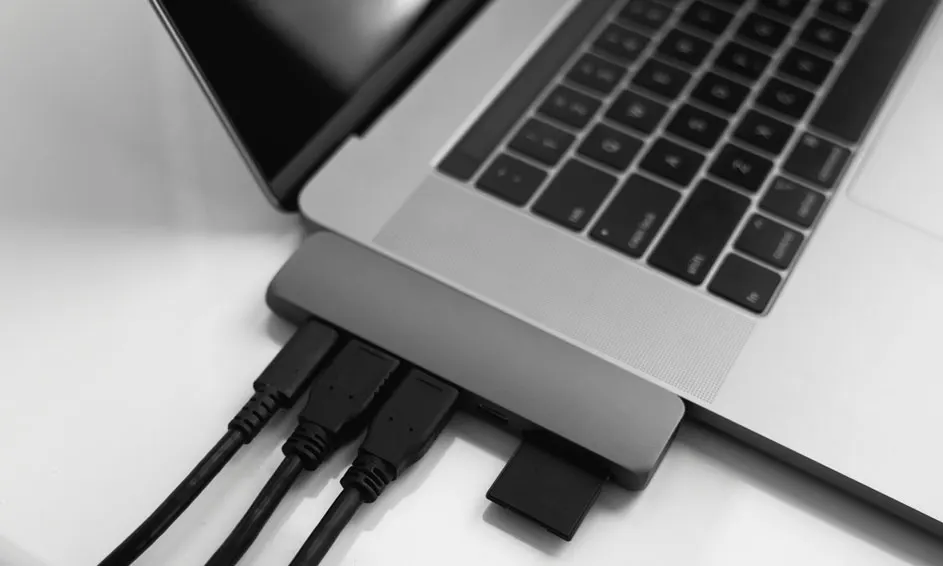 Ordinateur portable gris connecté à un concentrateur avec des câbles noirs et une carte mémoire sur une surface blanche.
