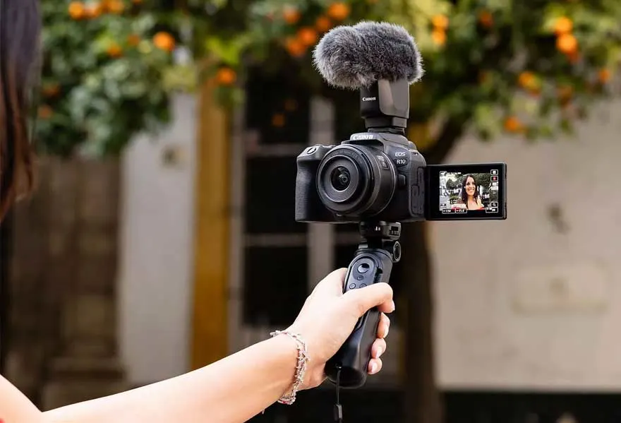 Een zwarte Canon EOS R10 camera met microfoon wordt vastgehouden, het scherm toont een video. Op de achtergrond: een boom met oranje vruchten.
