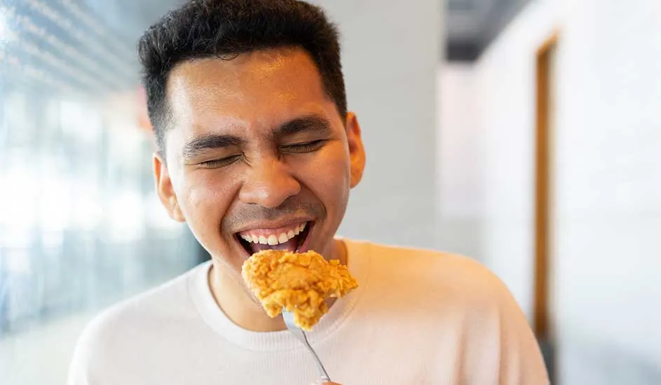 A person in a white jumper holds a fork with a bite of food, seated in a bright, modern space featuring glass and wood.
