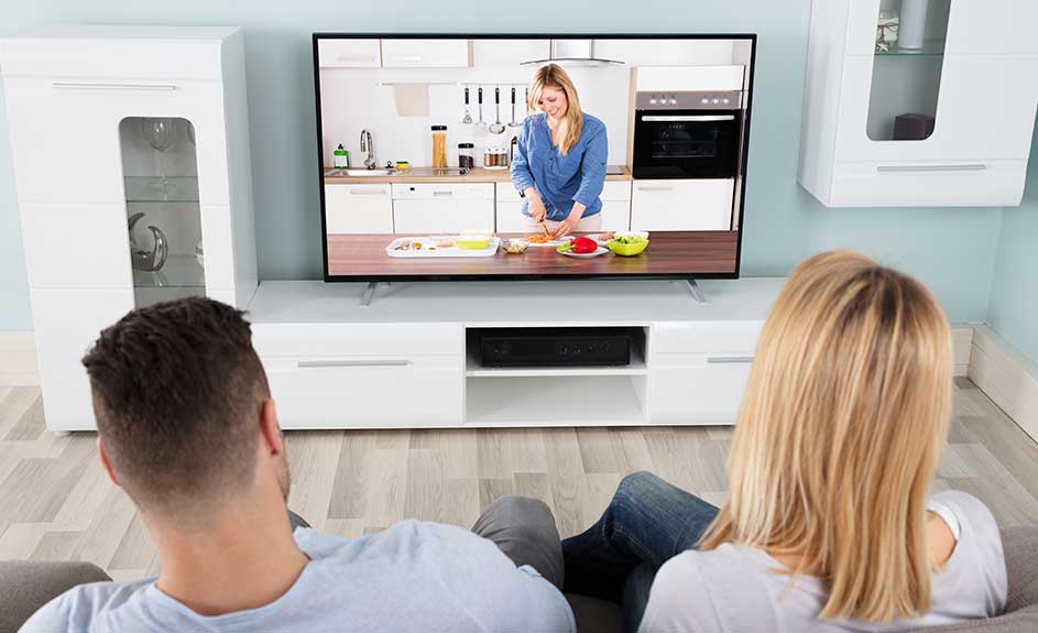 A TV shows a woman in a kitchen. Cabinets flank the TV on a white stand, viewed from behind a couch.