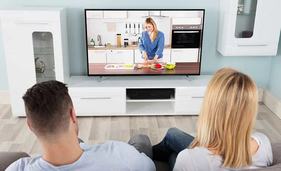 A TV shows a woman in a kitchen. Cabinets flank the TV on a white stand, viewed from behind a couch.