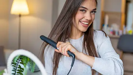 Interior shot of a black flat iron being used on long, brown hair. Lamp and plant in the background.