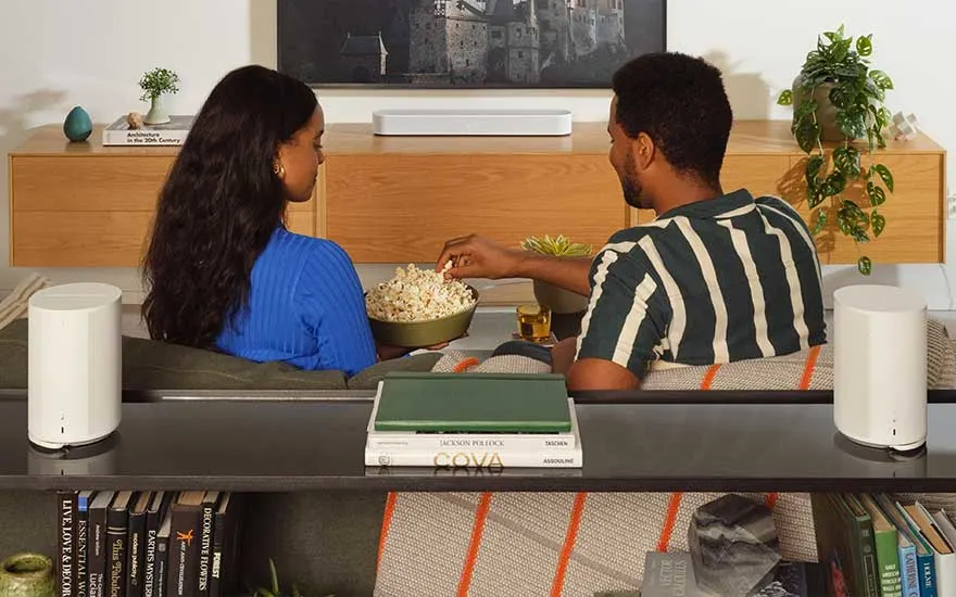 Woonkamer met tv, houten kast, planten en decoratie. Een kom popcorn staat centraal, omringd door boeken en witte speakers.