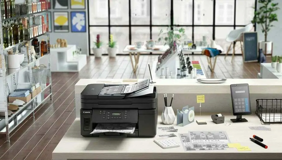 Une imprimante noire sur une table blanche avec des papiers, près d'une fenêtre et d'étagères dans une pièce lumineuse.