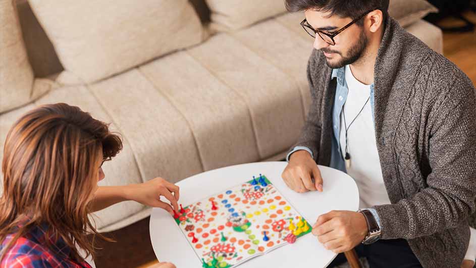 Twee personen spelen een bordspel op een witte tafel. Het spel heeft kleurrijke pionnen en een parcours. Een beige sofa is zichtbaar op de achtergrond.