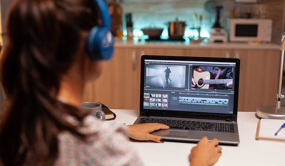 Een laptop toont videobewerkingssoftware met clips van een persoon en een gitaar. Er staat een mok en notitieboek op een wit bureau.