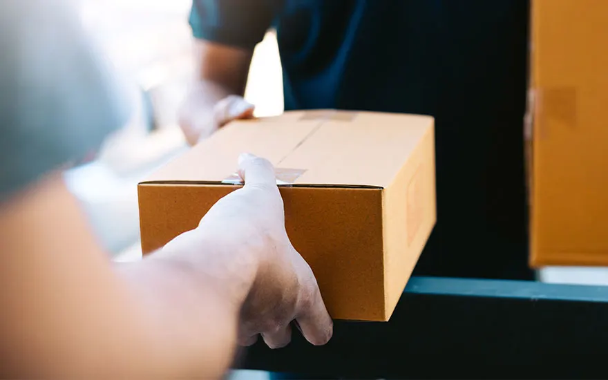 Livraison : Mains tenant une boîte en carton marron. Une autre boîte et un vêtement foncé sont visibles en arrière-plan.