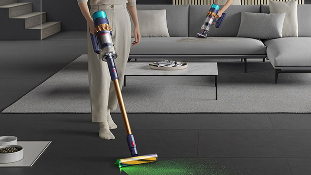 Un aspirateur nettoie un sol foncé; un autre est sur un sofa gris. Une table basse blanche est au centre.