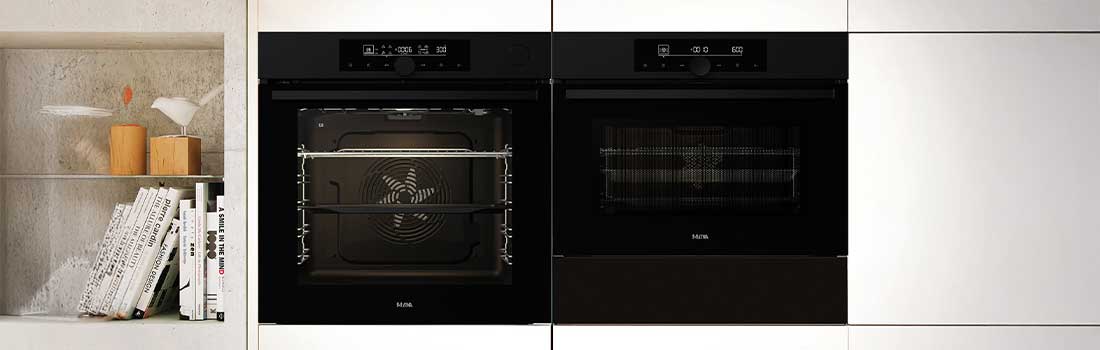 Two black ovens are set in white cabinets. A shelf shows books and decor.