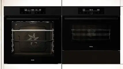 Two black ovens are set in white cabinets. A shelf shows books and decor.