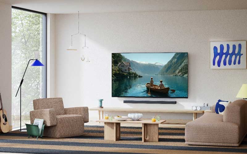 Salon lumineux avec une télévision murale montrant des personnes dans une barque sur un lac de montagne, avec un canapé, un fauteuil et de grandes fenêtres.
