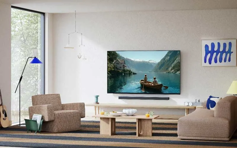 Salon lumineux avec une télévision murale montrant des personnes dans une barque sur un lac de montagne, avec un canapé, un fauteuil et de grandes fenêtres.