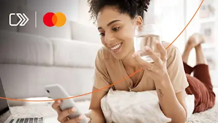 Een laptop en smartphone op een vloerkleed, met logo's in de hoek, een glas water in de hand, en een bank op de achtergrond.