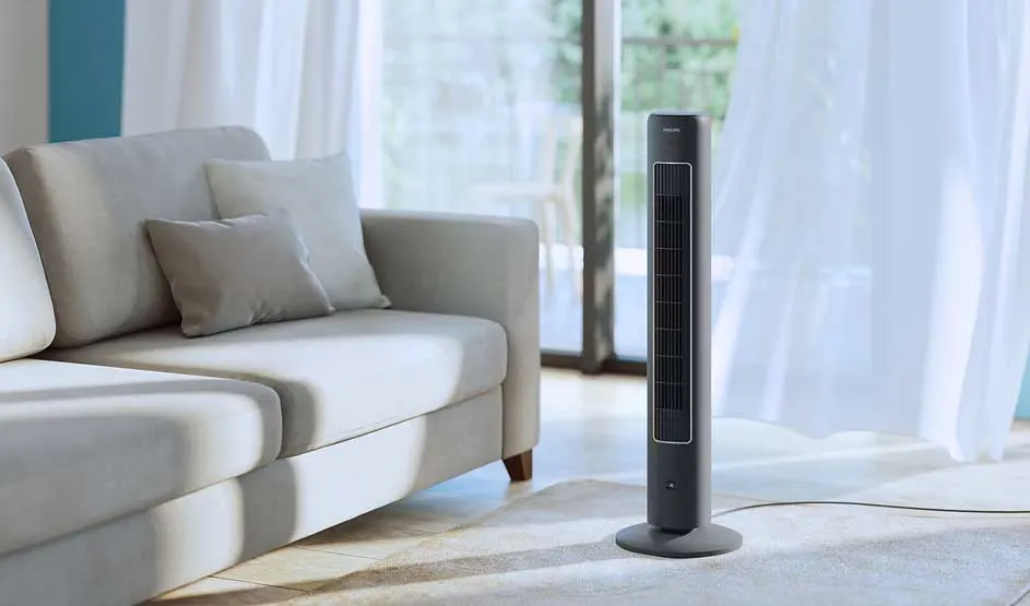 Een slanke torenventilator staat naast een lichte bank in een rustige, zonnige woonkamer met doorschijnende gordijnen.