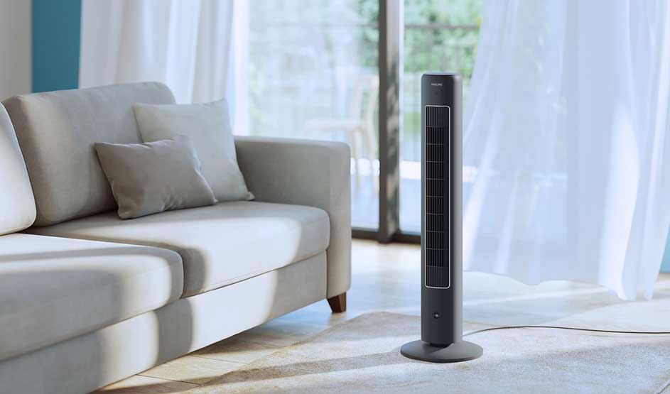 A slim tower fan stands beside a light sofa in a calm, sunlit living room with sheer curtains.