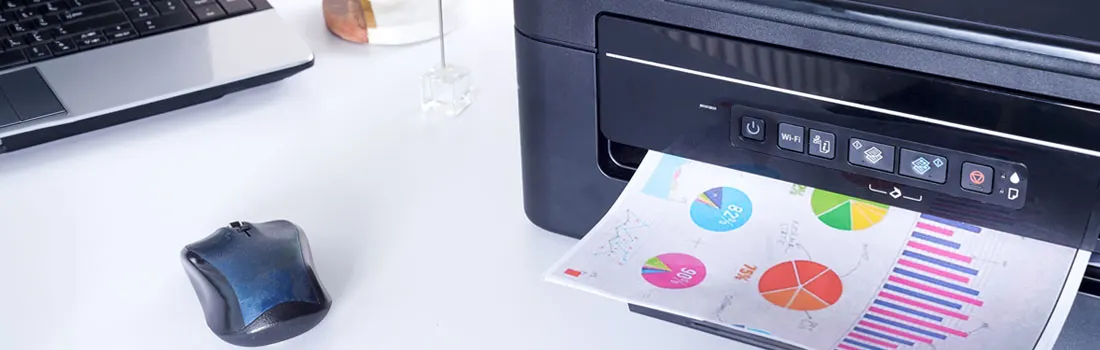 A printer outputs a page with colorful charts. A laptop and mouse are on the white desk.