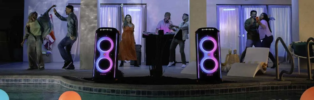 Outdoor party scene: Two light-up speakers flank a DJ setup by a pool. Floats drift in the water. Buildings stand in the background.