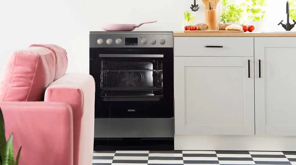 Een keuken met een roze zetel, een zwart-zilveren oven en witte kasten. Een roze pan ligt op de oven. De vloer heeft een zwart-wit dambordpatroon.