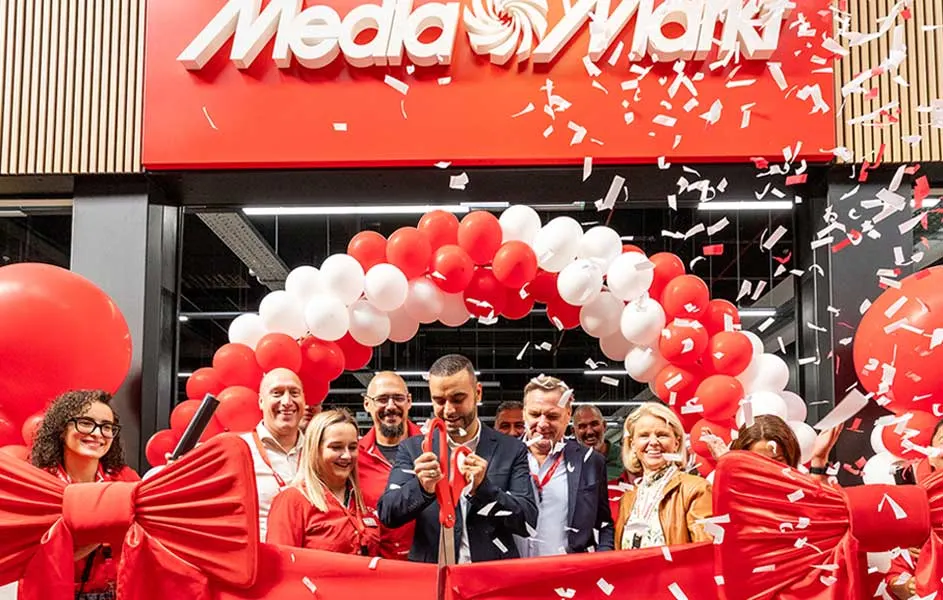 Voor een MediaMarkt-winkel staan lachende medewerkers en bezoekers onder een boog van rode en witte ballonnen, terwijl confetti door de lucht vliegt.