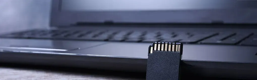 Une carte SD noire se trouve devant un ordinateur portable gris. Les broches dorées de la carte sont visibles.