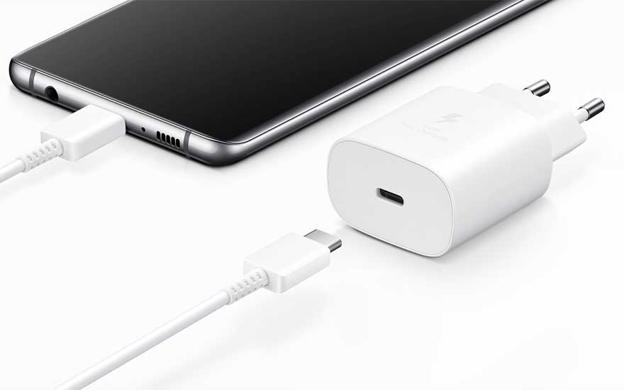 Un smartphone noir est en charge avec un câble USB-C blanc et un adaptateur secteur blanc sur une surface blanche.