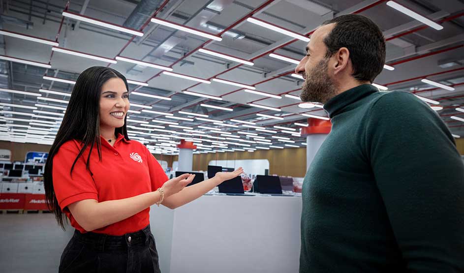 Een winkelmedewerkster in rood praat glimlachend met een klant in een grote, heldere elektronicawinkel.