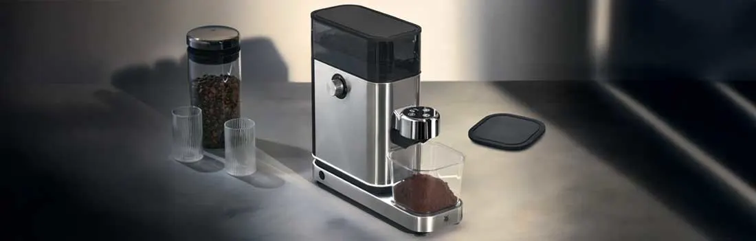 Een compacte koffiemolen staat op een aanrecht, naast twee maatbekers en een pot koffiebonen. Het apparaat oogt modern en glanzend.
