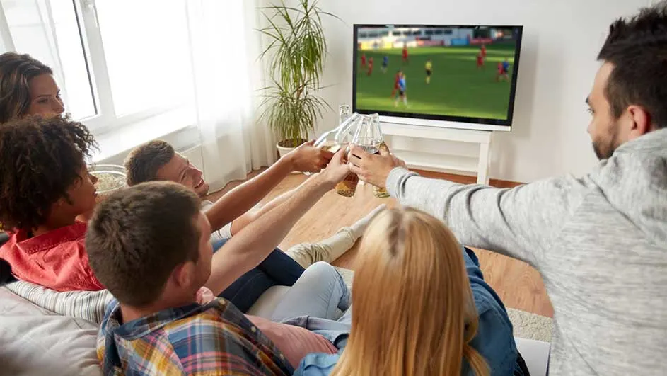 Een groep mensen zit op een bank met bier en kijkt naar voetbal op tv in een lichte woonkamer.