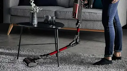 Un aspirateur noir et rouge nettoie un tapis gris. Une table noire avec vase et verres est près d'un canapé gris.
