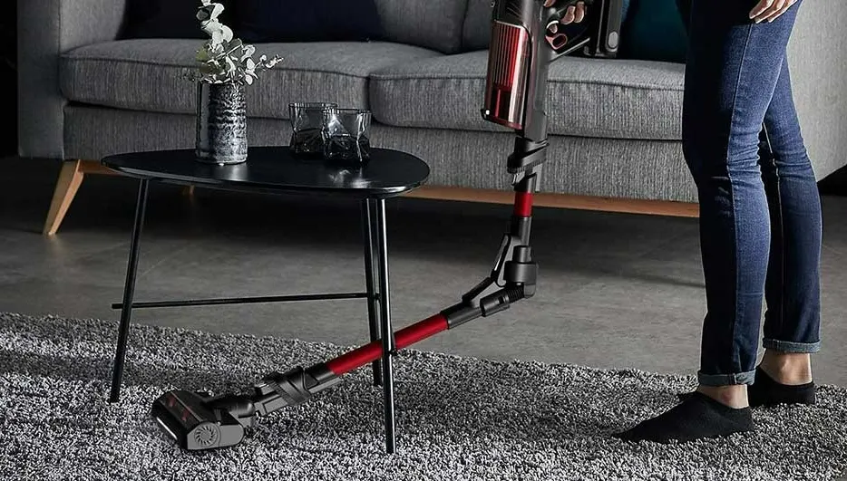 Un aspirateur noir et rouge nettoie un tapis gris. Une table noire avec vase et verres est près d'un canapé gris.