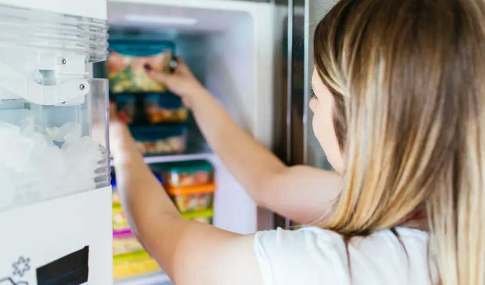 Une personne ouvre un frigo blanc rempli de boîtes colorées. Un distributeur de glace est visible.