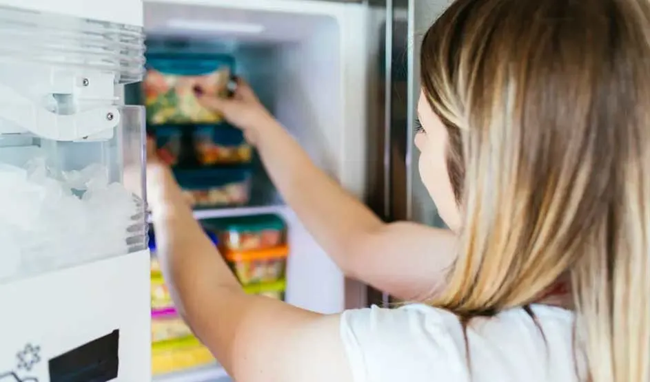Une personne ouvre un frigo blanc rempli de boîtes colorées. Un distributeur de glace est visible.
