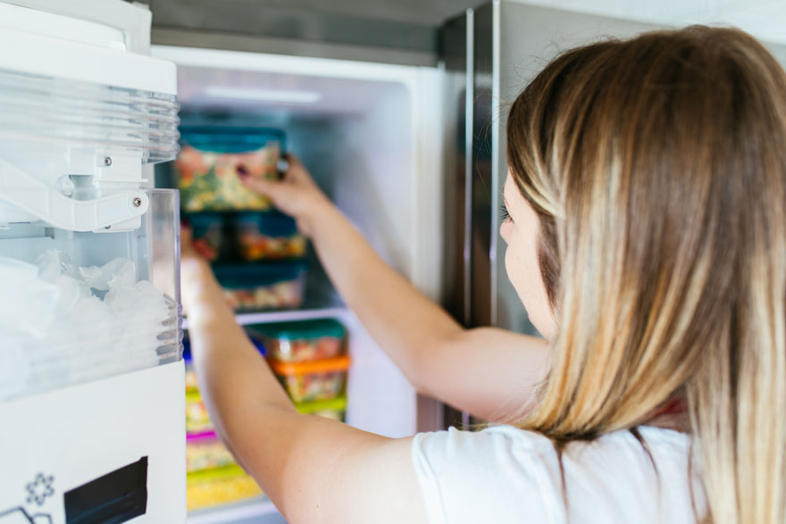 Une personne ouvre un frigo blanc rempli de boîtes colorées. Un distributeur de glace est visible.
