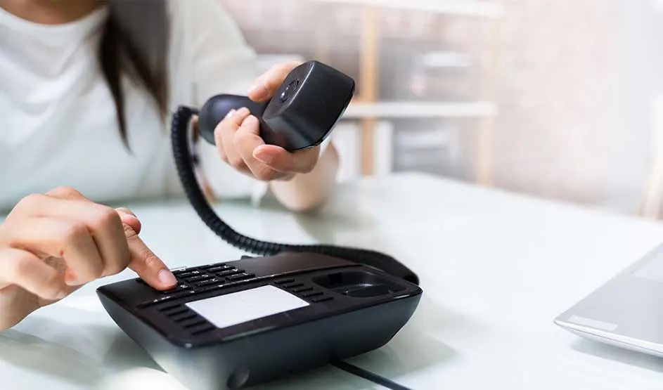Une personne utilise un téléphone fixe sur un bureau, tient le combiné et appuie sur les touches près d’un ordinateur portable.