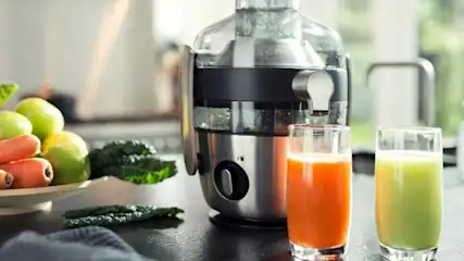 Un presse-agrumes argenté et noir à côté de verres de jus orange et vert. Assiette de carottes et de pommes vertes en arrière-plan.