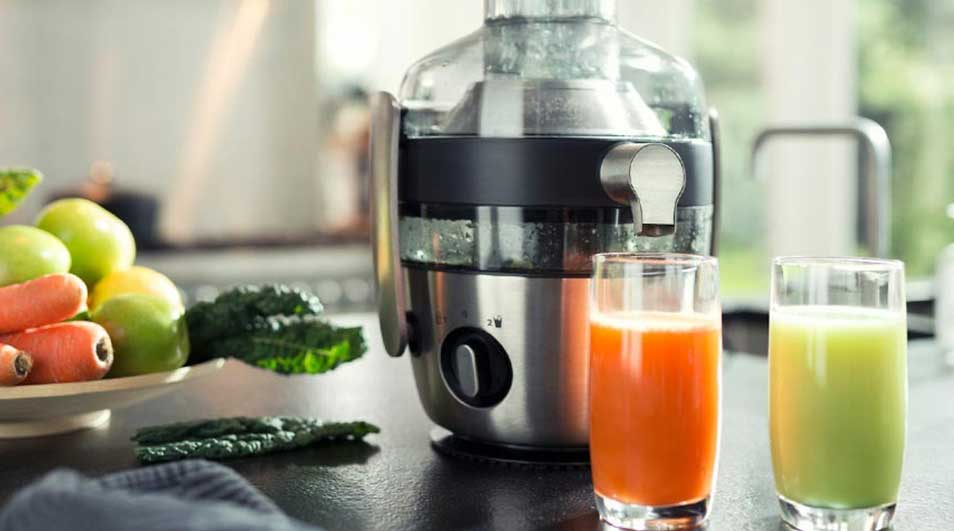Een zilverkleurige slowjuicer naast glazen oranje en groene sap. Op een bord liggen wortelen, groene appels en boerenkool.