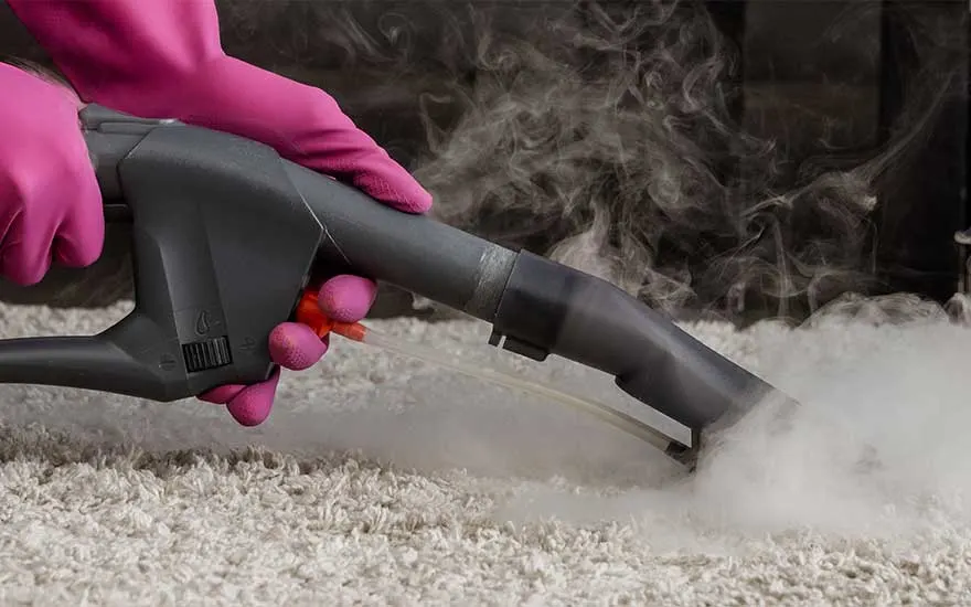 Une machine grise nettoie un tapis blanc, dégageant de la vapeur. Des gants roses tiennent la machine. Arrière-plan sombre.