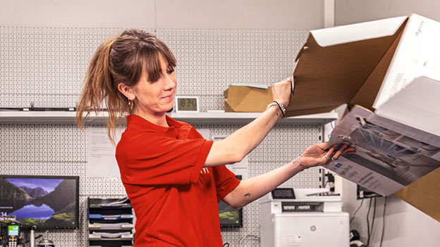 Une personne en T-shirt rouge sort un objet d'une boîte en carton dans un bureau. À l'arrière-plan, un moniteur et une imprimante.