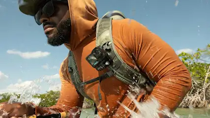 Iemand staat in het water met een oranje hoodie en een groene rugzak met camera. Op de achtergrond blauwe lucht en groene begroeiing.