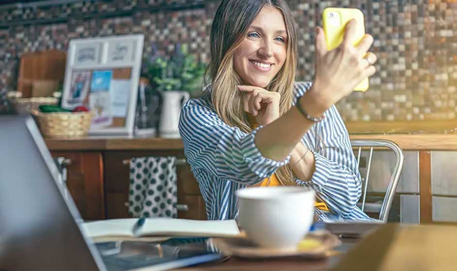 Een glimlachende persoon zit in een café en maakt een selfie met een gele telefoon, naast een kop koffie, laptop en notitieboek.